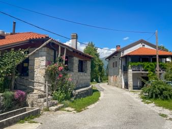 Narrow paved road through Robidisce with traditional stone houses featuring red tile roofs and flowering plants under a clear blue sky.