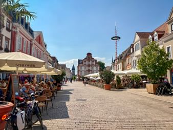 Kopfsteinpflaster-Fußgängerzone Maximilianstraße in Speyer mit Straßencafés, bunten historischen Gebäuden und einem Maibaum unter blauem Himmel.