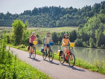 Drei Radfahrer fahren auf asphaltiertem Weg entlang der Mur mit bewaldeten Hügeln im Hintergrund. Sonniger Tag mit ruhigem Wasser.