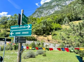 Directional cycling signs pointing to Tarvisio-Austria and Udine-Grado in the Kanaltal valley, with mountains and colorful flags in background.