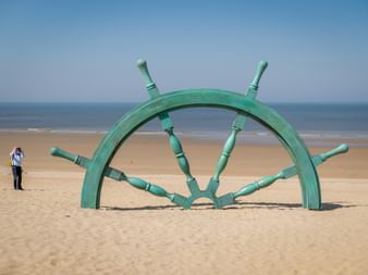 Großes grünes Steuerrad-Denkmal, halb im Sand vergraben am Strand von Westende mit Person daneben. Blauer Himmel und Meer im Hintergrund.