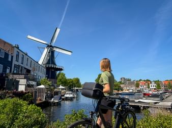 Female cyclist with bike viewing De Adriaan windmill across canal in Haarlem. Historic buildings and boats line the waterway under blue sky.