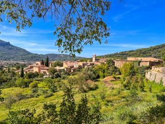 Biniaraix village with stone houses and church tower, surrounded by green fields and mountains under blue sky in Mallorca.