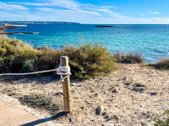 Sandy coastal path with rope fence along turquoise Mediterranean Sea in Porto Colom, Mallorca. Green vegetation and small islands visible.