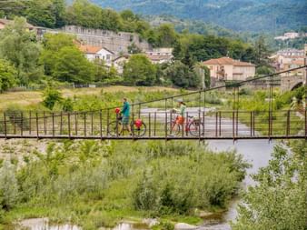 Zwei Radfahrer mit Rädern überqueren eine Metallbrücke über einen Fluss in Millesimo, Piemont. Dorfgebäude und grüne Hügel im Hintergrund.