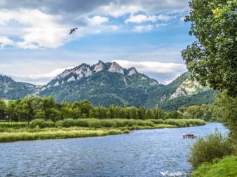 Dunajec River flowing through green valley with Trzy Korony Mountains in background. A raft with passengers floats on the water under cloudy sky.