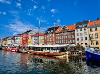Historic Nyhavn harbor in Copenhagen with colorful townhouses, wooden boats moored along the canal, and outdoor cafes under a blue sky.