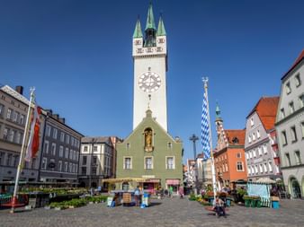 Straubinger Marktplatz mit weißem Stadtturm, grünem Gebäude, bayerischer Fahne und bunten historischen Häusern unter blauem Himmel.