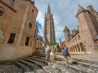 Drei Personen steigen Steinstufen im Innenhof des Gruuthuse-Palasts in Brügge mit gotischem Kirchturm und historischen Backsteingebäuden.
