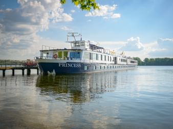 White river cruise ship MS Princess docked at a wooden pier on calm water under blue sky with white clouds and green trees visible.