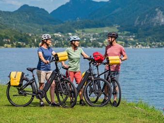 Three cyclists with e-bikes and yellow panniers standing on grass by Lake Wolfgang. Mountains and village visible in background.