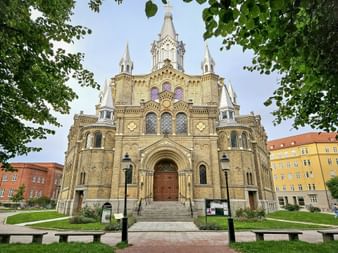 Yellow brick church with ornate facade and multiple spires in Riga, framed by green trees. Colorful buildings flank the church on both sides.