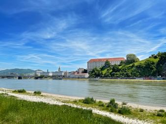 Blick auf das Linzer Schlossmuseum auf einem grünen Hügel über der Donau. Historische Gebäude und eine Brücke sind am Ufer unter blauem Himmel sichtbar.