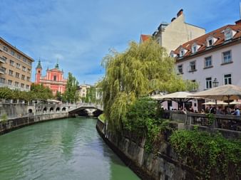 Grüner Fluss durch Ljubljana mit rosa Kirche, Steinbrücke, Weide und Flusscafés unter blauem Himmel.