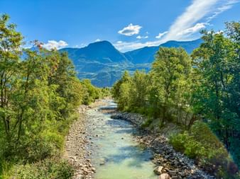 Shallow river with rocky bed flowing between green trees toward mountains under blue sky near Brixen, South Tyrol.