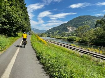 Radfahrer in gelbem Trikot auf asphaltiertem Radweg neben Bahngleisen, mit grünen Bergen und blauem Himmel in österreichischer Landschaft.