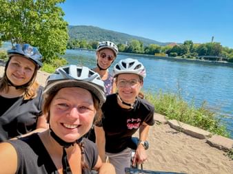 Vier Radfahrerinnen mit Helmen machen ein Selfie am Sandstrand am Neckar in Heidelberg, mit Hügeln und blauem Himmel im Hintergrund.