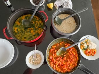 Multiple pots and pans on dark surface containing colorful vegetable dishes including spinach curry, rice, and diced carrots with spoons.