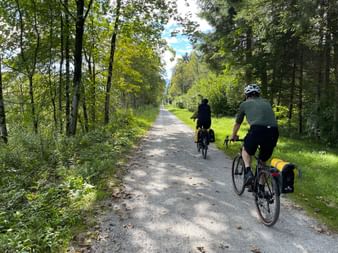 Two cyclists with panniers riding on a gravel path through a lush green forest in Bohemia, Czech Republic.