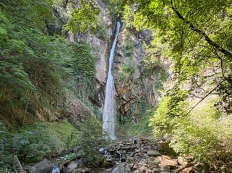 Tall waterfall cascading down rocky cliff surrounded by lush green vegetation along the Brandis Waalweg near Meran, with rocky stream bed below.