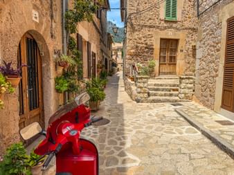 Roter Vespa-Roller in enger Steingasse in Valldemossa, Mallorca. Steingebäude mit Holzläden säumen die Straße mit Topfpflanzen.