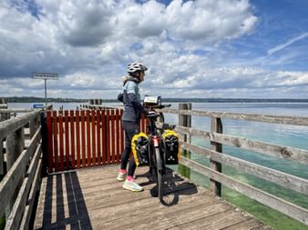 Female cyclist with helmet and touring bike with yellow panniers standing on wooden pier in Ambach, overlooking Lake Starnberg under cloudy sky.