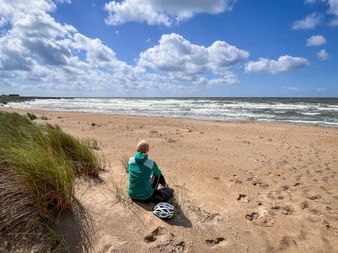 Cyclist in green jacket sitting on sandy beach with bicycle helmet nearby, overlooking North Sea waves under cloudy blue sky.