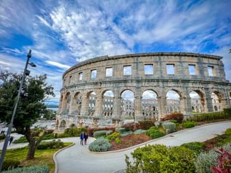 Antikes römisches Amphitheater Arena von Pula in Istrien mit Steinbögen und angelegten Gärten unter blauem Himmel mit weißen Wolken.