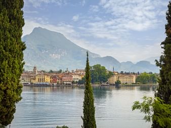 Blick auf Riva del Garda am Gardasee, eingerahmt von Zypressen. Bunte Gebäude säumen das Ufer mit Bergen im Hintergrund.
