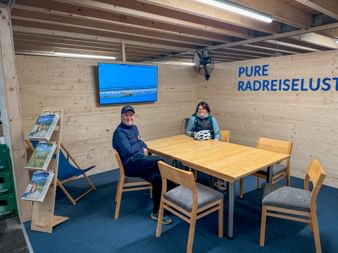 Zwei Radfahrer sitzen an Holztisch in modernem Besucherzentrum mit sichtbaren Deckenbalken, blauer Wandbildschirm und 'PURE RADLEISELUST' Schild.