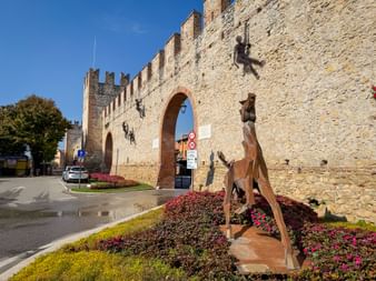 Medieval crenellated city wall in Soave with arched gateway. Metal sculptures of climbing figures on wall and flower beds in foreground.