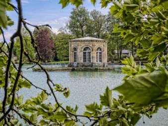 Classical stone pavilion with arched windows on a pond in Fontainebleau park, framed by green foliage and surrounded by trees under blue sky.