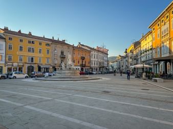 Wide square with fountain in center, surrounded by colorful historic buildings in Gorizia. Clear blue sky and mountains visible in background.