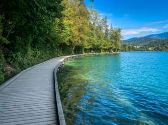 Geschwungener Holzsteg am Bleder See mit türkisfarbenem Wasser, umgeben von grünen Bäumen und Bergen im Hintergrund unter blauem Himmel.