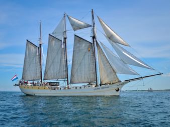 Three-masted sailing ship Mare Frisium with white sails unfurled on calm blue waters under a clear sky, with Dutch flag visible.