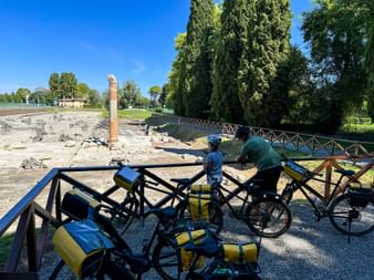 Two cyclists with yellow panniers viewing ancient Roman ruins in Aquileia. Stone columns and excavations visible under blue sky with cypress trees.