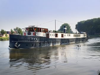 MS Magnifique II river cruise ship with blue hull and white superstructure sailing on calm water. Trees and buildings visible on shore.