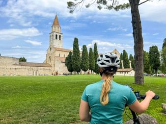 Female cyclist with white helmet viewing the Basilica di Santa Maria Assunta in Aquileia across a green lawn with cypress trees.