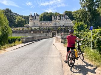 Radfahrer mit Helm und gelben Taschen auf Brücke vor Schloss Ussé, einem Renaissance-Schloss umgeben von Bäumen an der Loire.