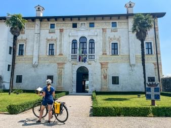 Radfahrerin mit Tourenrad und Karte vor einem weißen historischen Palazzo mit verzierter Fassade, flankiert von Palmen in Friaul.