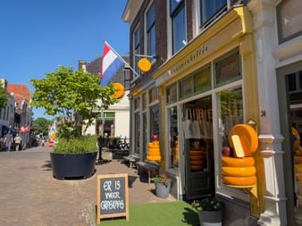 Traditional cheese shop in Gouda with yellow facade displaying stacked cheese wheels. Dutch flag flies above, chalkboard sign reads 'er is weer graskaas'.