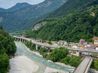 Erhöhte Betonbrücke über einen türkisfarbenen Fluss in Dogna, eingebettet in ein grünes Bergtal zwischen Tarvis und Gemona auf der Bled-Venedig-Route.