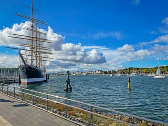 Large historic sailing ship with multiple masts moored at Travemünde harbor entrance, with marina and coastal buildings in background.