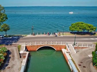 Rote Ziegelbrücke über grünem Kanal am Lido di Venezia. Radfahrer auf Promenade zwischen Kanal und blauer Adria mit weißem Boot in der Ferne.
