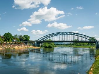 Stahlbogenbrücke über die Weser in Rinteln mit Sandstrand am linken Ufer, Bäumen und weißen Wolken am blauen Himmel.