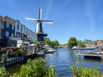 Historische Windmühle De Adriaan an einem Kanal in Haarlem mit Booten am Ufer und traditionellen holländischen Häusern unter blauem Himmel.
