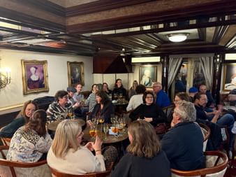 Gruppe speist in elegantem Schiffsrestaurant mit dunklen Holzbalken, klassischen Porträts und runden Tischen. Menschen genießen Getränke.