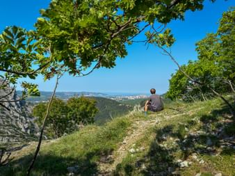 Cyclist sitting on grassy hillside path in Val Rosandra under trees, overlooking distant view of Trieste and the Adriatic Sea under blue sky.