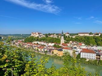 Panoramablick auf Burghausen mit Burganlage auf einem Hügel, Kirchturm und bunten Gebäuden entlang des Flusses, eingerahmt von grünem Laub.