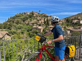 Cyclist with red bike and yellow panniers at viewpoint overlooking the hillside village of Deià in Mallorca under blue sky.